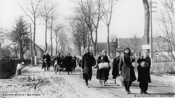 Historyczne czarno-białe zdjęcie przedstawia grupę przesiedleńców, głównie kobiet, niosących torby i walizki przez wieś w Prusach Wschodnich, prawdopodobnie wiosną 1946 r.