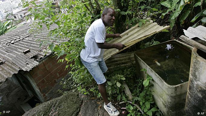 Homem inspeciona caixa d'água nos fundos do quintal de sua casa, cercada pela mata em um morro do Rio de Janeiro.
