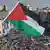 Eine Hand schwenkt eine palästinensische Flagge, im Hintergrund sind auf einem Platz und auf umliegenden Hausdächern in Ramallah zahlreiche Demonstrationen zu sehen