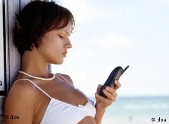 Ein junge Frau in Strandkleidung schickt mit ihrem Mobiltelefon eine SMS-Nachricht (Short Message Service) ab.
