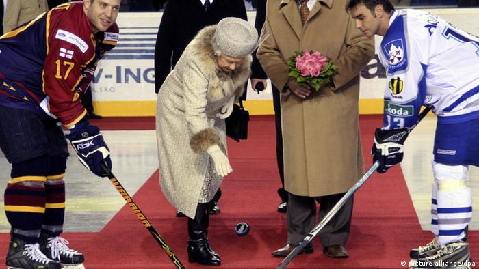 Queen Elizabeth II puts a puck on a hockey rink with two hockey players standing nearby.