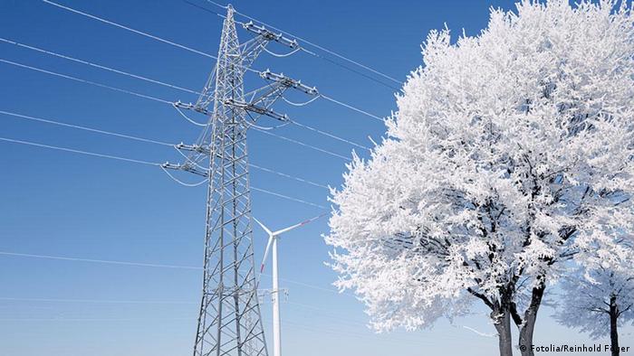 An electricity pylon and a tree covered in snow shown in front of a windmill