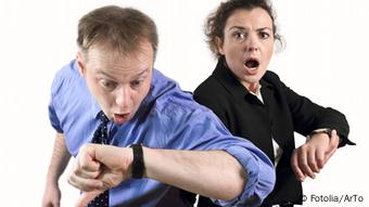 Ein Mann in Hemd und Krawatte (vorne ) sowie eine Frau im Businesskostüm schauen entsetzt auf ihre Armbanduhren