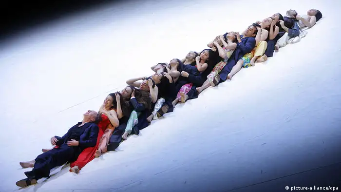 Tänzer sitzen hintereinander und ineinander verschränkt auf der Bühne und bilden eine Linie.
(c) dpa - Bildfunk