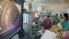 Tela de TV mostra interior de intestino, com médicos ao fundo realizando colonoscopia em paciente deitado