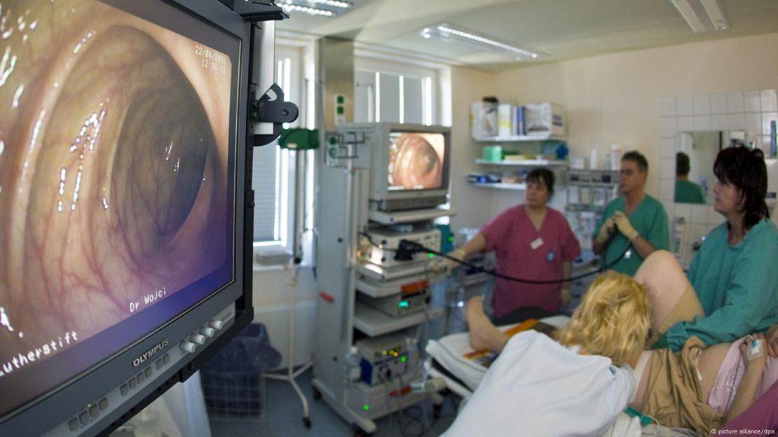 Tela de TV mostra interior de intestino, com médicos ao fundo realizando colonoscopia em paciente deitado