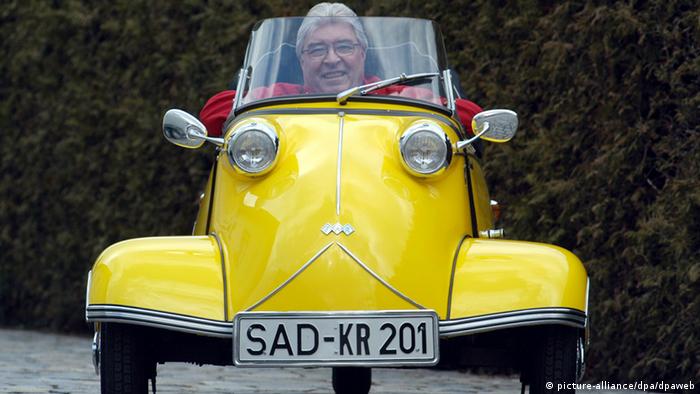 Ein lächelnder Mann sitzt in einem kleinen, gelben Oldtimer