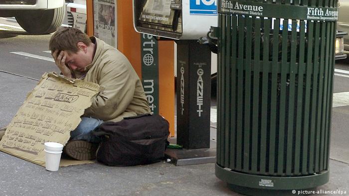 Ein Bettler mit einem Pappschild und einem Pappbecher vor sich sitzt auf dem Bürgersteig neben einem Mülleimer auf dem Fifth Avenue steht (Foto: picture-alliance/dpa)