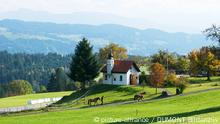 Eine Kapelle in bergigem Land, davor zwei Menschen, die Pferde führen.