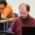 Homem de barba e óculos sentado diante de computador, com outro homem sentado ao computador numa mesa atrás dele