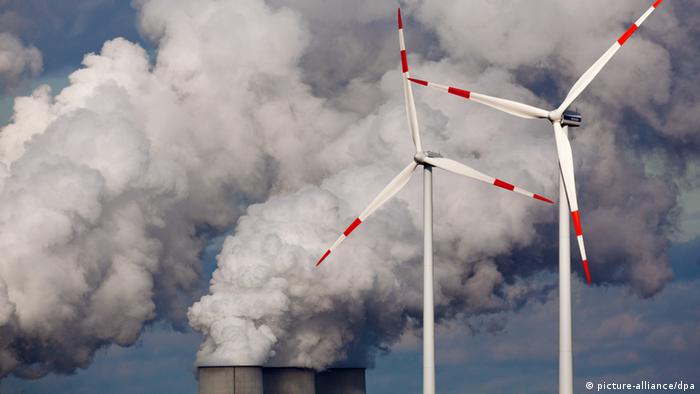 Two wind turbines against a backdrop of plumes of smoke billowing from factory chimneys