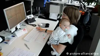 Symbolbild - Eine junge Mutter sitzt mit ihrem Baby an ihrem Schreibtisch, auf dem der Monitor eines Computers steht. Foto: Tim Brakemeier