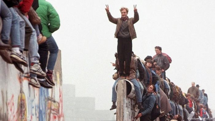 Ein Mann steht auf der Berliner Mauer, vor ihm klafft eine Lücke, er formt mit seinen Fingern das Victory-Zeichen