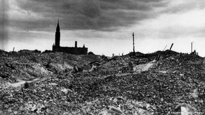 Auf einem schwarz-weiß-Foto ist eine Trümmerwüste aus Steinen zu sehen, um Hintergrund erkennt man ein Gebäude mit Turm, es könnte eine Kirche sein