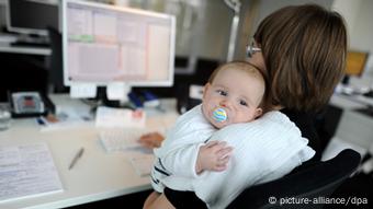 Eine junge Mutter sitzt mit ihrem Baby an ihrem Schreibtisch, auf dem der Monitor eines Computers steht. (Foto: Tim Brakemeier / picture alliance / dpa)