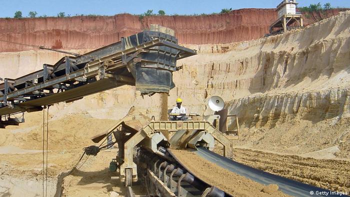 In einem Tagebau in Togo wird Phosphorgestein abgebaut. Von einem Kran fällt Sand auf ein Förderband. Ein Arbeiter beobachtet den Vorgang. Die Erde über dem Tagebau ist rot-braun. 
