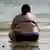 Eine sehr übergewichtige Frau sitzt im Bikini am Strand und schaut auf das Meer hinaus.