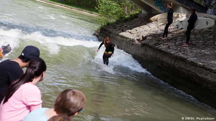 Eine junge Frau surft auf dem Fluss Isar. Drei Zuschauer sehen ihr zu.