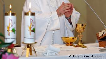Ein Priester steht am Altar, rechts neben ihm stehen zwei brennende Kerzen. Er hat die Hände gefaltet, vor ihm steht ein Messkelch