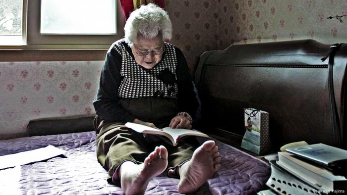 Lee Ok-Seon in ihrem Zimmer mit einem Buch auf dem Schoß auf dem Bett (Foto: Tsukasa Yajima)