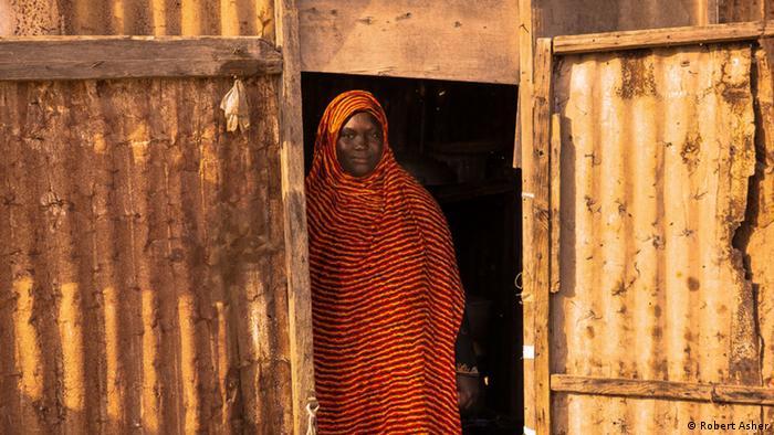 A woman stands in a doorway to a makeshift shack