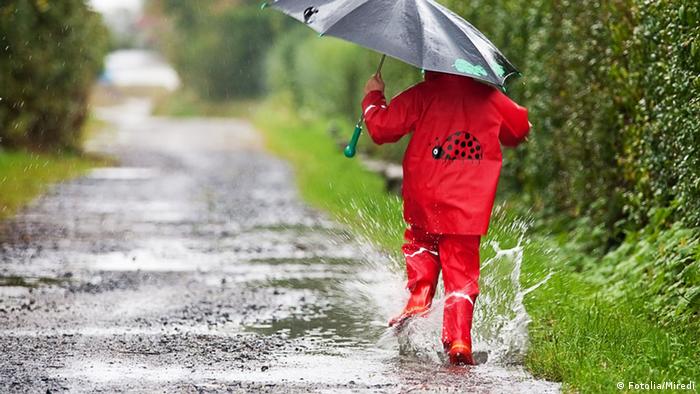 A child in a red rain suit and with black umbrella splashes water from a puddle.