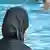 A woman wearing a black headscarf and robes looks away from the camera and toward a swimming pool where people are swimming