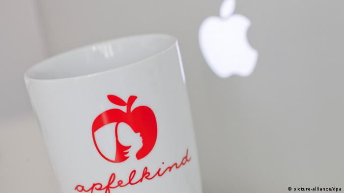 A white coffee mug emblazoned with a red, apple logo is compared against the backlit white Apple logo of a gray laptop computer. Photo: Rolf Vennenbernd
