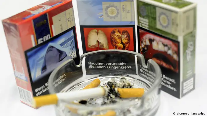 ARCHIV - Zigarettenschachteln mit einem möglichen einheitlichem Aussehen und Aufdrücken von Folgeschäden des Zigarettenkonsums stehen am 14.09.2012 auf der Eröffnungs-Pressekonferenz zur Tabak-Fachmesse inter-tabac in den Westfalenhallen in Dortmund hinter einem Aschenbecher mit einer qualmenden Zigarette. Foto: Jonas Güttler/dpa (zu dpa: Tabakrichtlinie/EU vom 08.10.2013) +++(c) dpa - Bildfunk+++