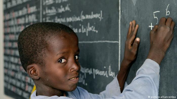 Ein kleiner afrikanischer Junge steht vor einer vollgeschriebenen Tafel mit einem Stück Kreide in der Hand. (Foto: picture-alliance/dpa)
