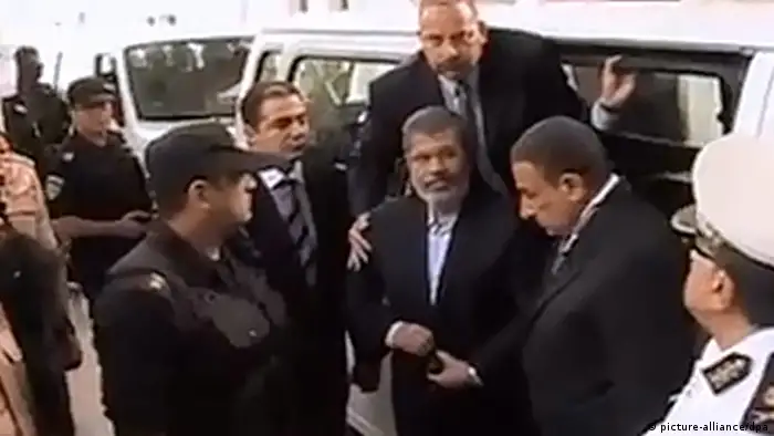 epa03935720 A grab made of video released by the Egyptian interior ministry showing ousted president Mohamed Morsi (C) arriving for his first trial session, in Cairo, Egypt, 04 November 2013. Egypt's toppled Islamist president, Mohammed Morsi, arrived on 04 November at the venue where his trial on charges of inciting the killing of opposition protesters is due to open. The so-called 'four-finger salute' has come to symbolize the lives lost during the dispersal of the Rabaa al-Adawiya protest camp by the Egyptian army. EPA/INTERIOR MINISTRY HANDOUT BEST QUALITY AVAILABLE HANDOUT EDITORIAL USE ONLY/NO SALES pixel