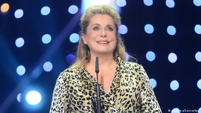 Catherine Deneuve stands a microphone onstage with lights shining behind her