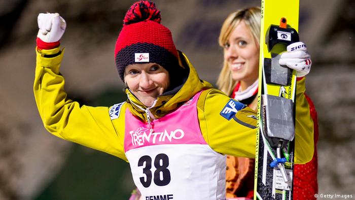 A woman holding a pair of skis and wearing a yellow ski suit raises her hands in triumph.
