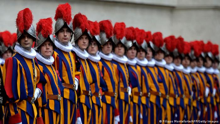 20 Soldaten der Päpstlichen Schweizergarde in ihrer blau-gelb-roten Uniform mit rotem Helmbusch in einer Reihe angetreten