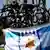 Holocaust survivors hold a flag of Greek-Jews Community and standing next to the Holocaust Memorial in Thessaloniki