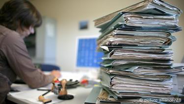 Куп документи едни врз други во преден план десно ставени на маса на која жена работи на компјутер Куп документи едни врз други во преден план десно ставени на маса на која жена работи на компјутер