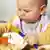 A baby sits at a high-chair eating from a plate of food