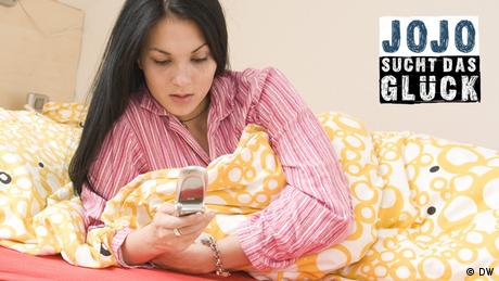 Eine Frau liegt in ihrem Bett und schaut auf ihr Handy. (DW)