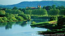 hinter einem langsam fließenden Fluss und versteckt von grünen Büschen und Bäumen erkennt man vor einem Hügel die Dächer einer Klosteranlage