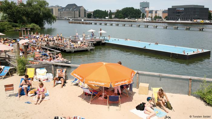 Badeschiff Berlin EINSCHRÄNKUNG
Im Hintergrund schwimmt das Badeschiff auf der Spree. Vorne entspannen Menschen auf dem Sandstrand. 