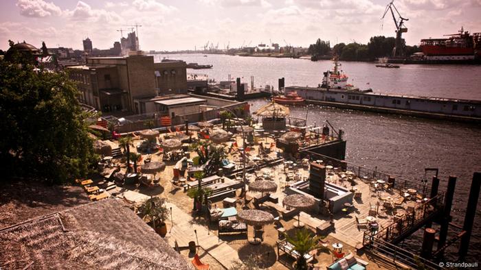 Strandpauli in Hamburg
Hafenkräne und die Elbe im Hintergrund. Mit Perspektive von oben das Gelände von Strandpauli mit Liegestühlen und Sonnenschirmen.