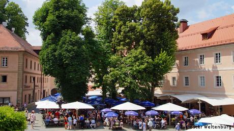 A biergarten with trees and blue umbrellas between peach-colored buildings