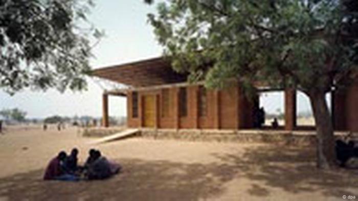  Burkina Faso, building in arid landscape, two trees and a group of people seated on the ground in front of it 