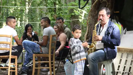 A young boy holds a microphone as a man plays the saxophone – the music is being played as part of a birthday party for one of the women at a Roma slum in Paris, France. The party goers sit laughing in the background. The community live in terrible conditions in the middle of a group of trees in the French capital with constant fear of being evicted from the site. 
