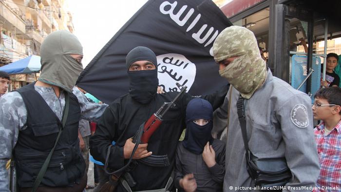 Maskierte al-Kaida-Kämpfer mit einer Fahne im Hintergrund und Maschinengewehren im Arme haben einen Jungen in ihrer Mitte. Auch er ist maskiert. Um sie verum stehen neugierige Jungen und Männer. (Foto: picture-alliance/ZUMA Press/Medyan Dairieh) 