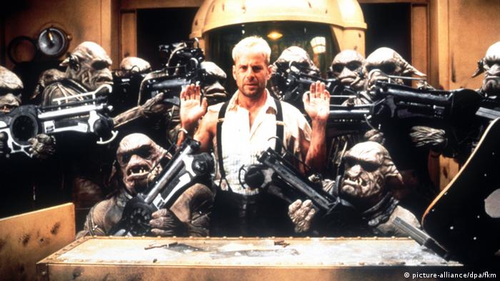 Film still The Fifth Element, man stands behind a table, arms raised, surrounded by heavily armed aliens