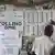 Two men point to voting lists hung up on a wall next to a poster that reads 'polling zone'