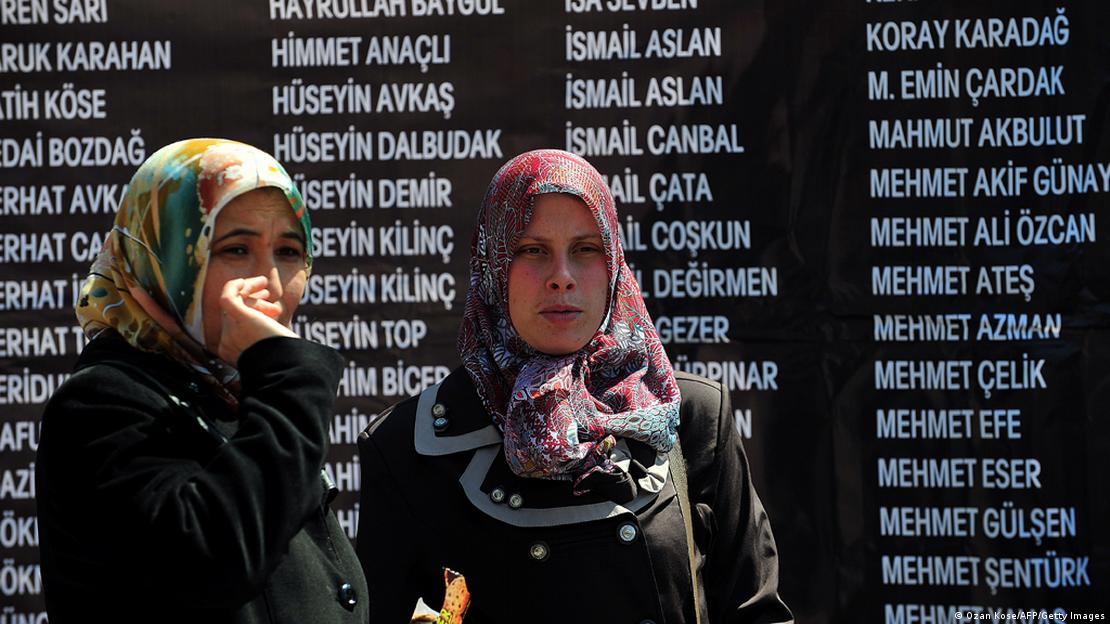 Soma'da 2015 yılında meydana gelen maden kazasında hayatını kaybedenlerin anısına düzenlenen törende iki kadın ağlarken. Arkalarında ölen işçilerin isimleri yazıyor 