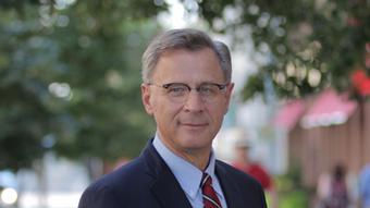 A middle-aged man wearing a dark suit and tie, and glasses with half a rim