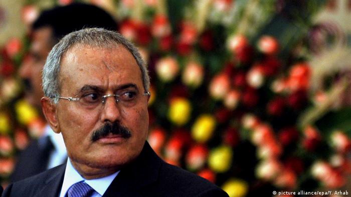 Former President Saleh looks off into the distance against a blurred background of red, yellow and green